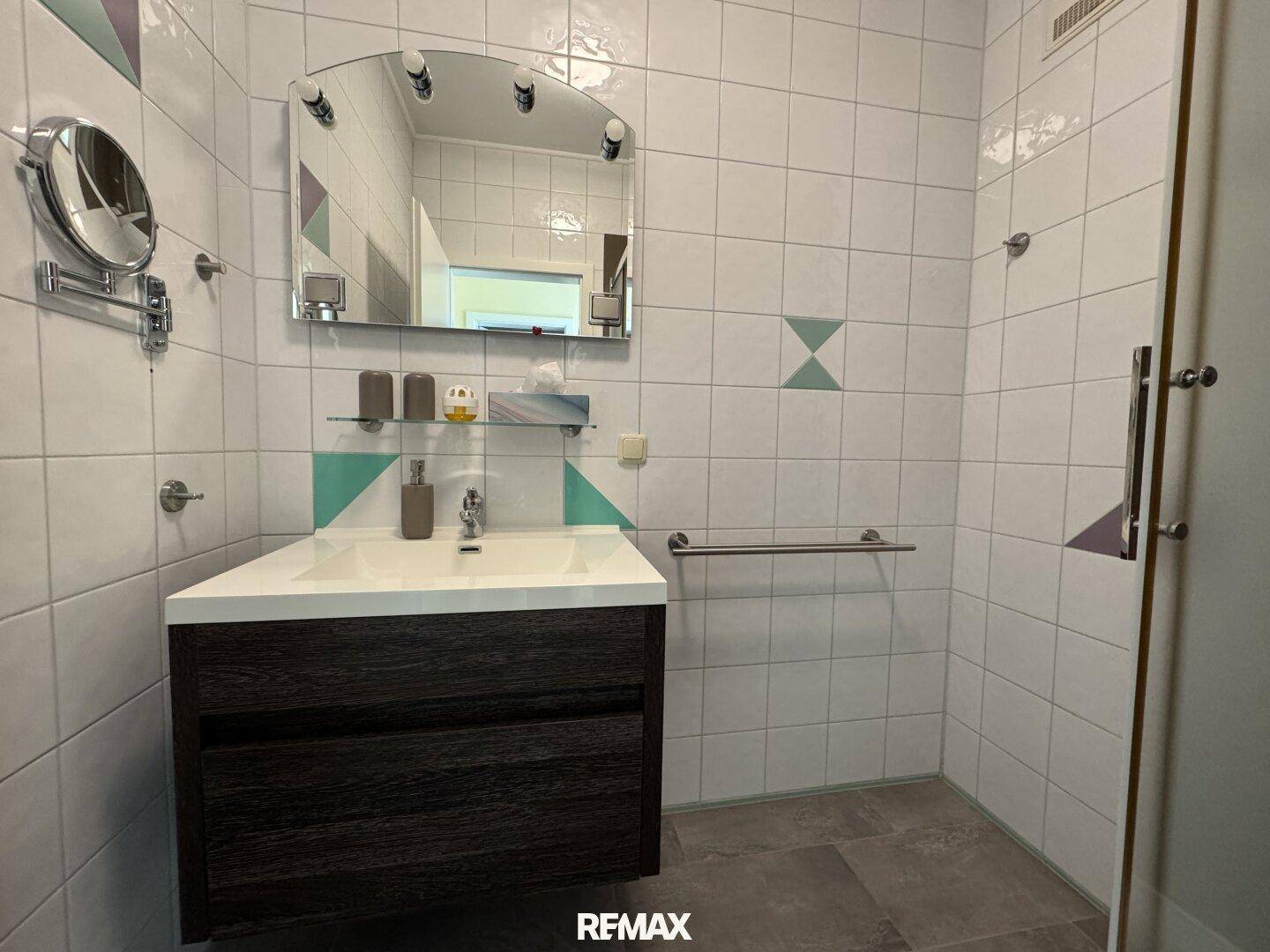 Modernes Badezimmer mit Waschtisch, Spiegel und Duschbereich, ausgestattet mit Haltegriff.