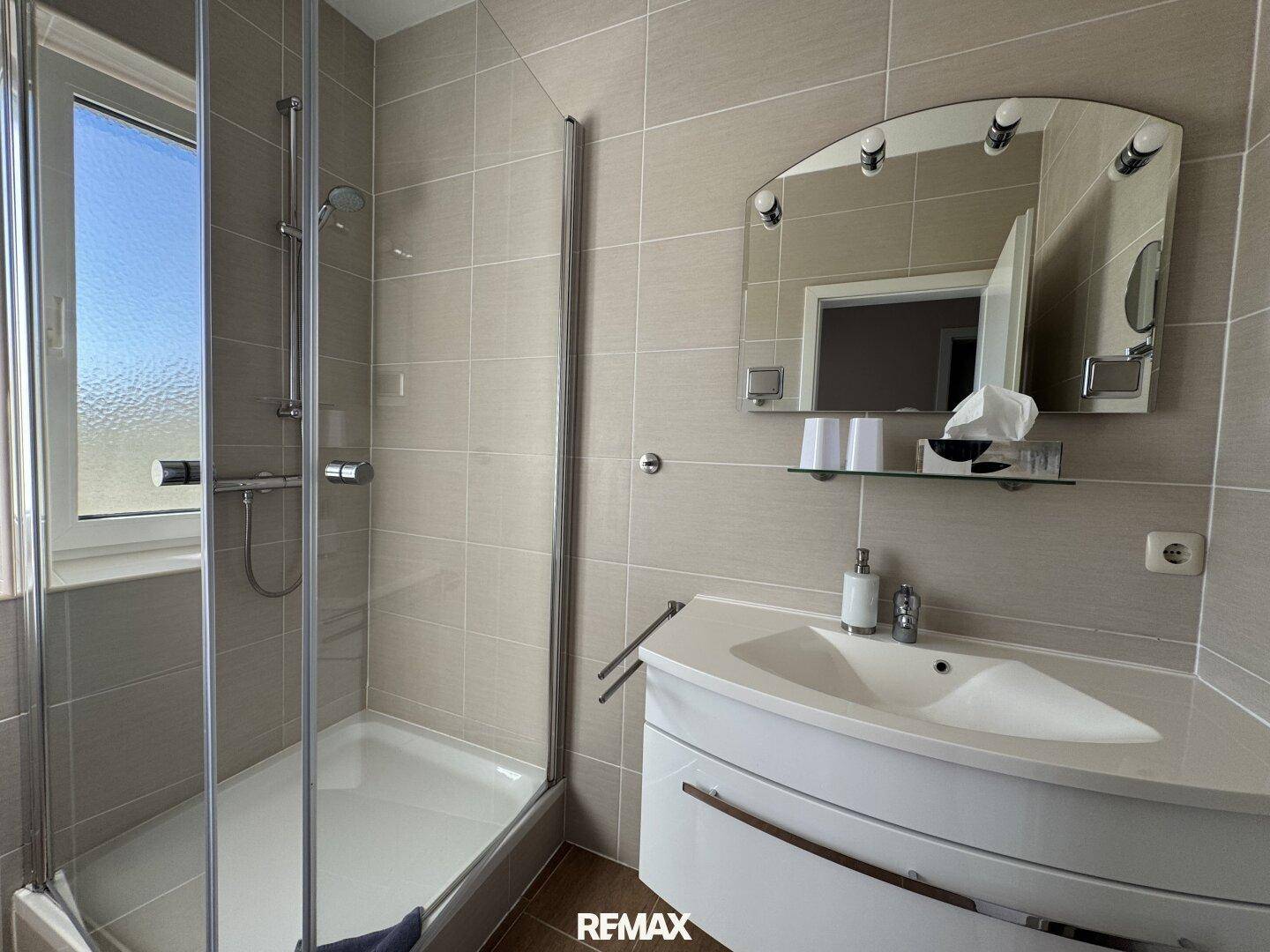 Modernes Badezimmer mit Eckwaschtisch, Spiegel und Duschkabine, in beigen Fliesen gehalten.