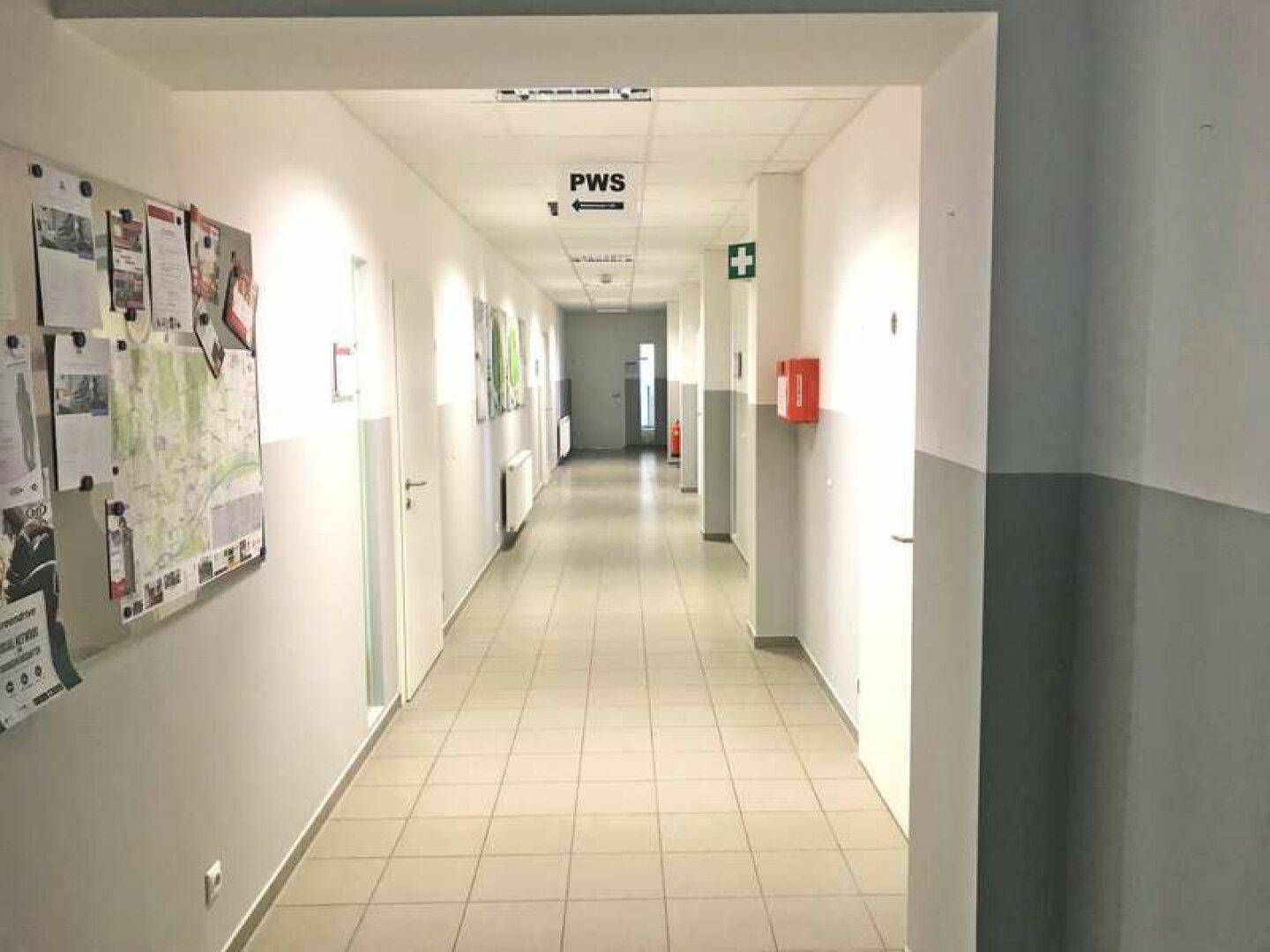 Langer, gut beleuchteter Flur mit Fliesenboden, Pinnwänden und Türen zu verschiedenen Räumen.