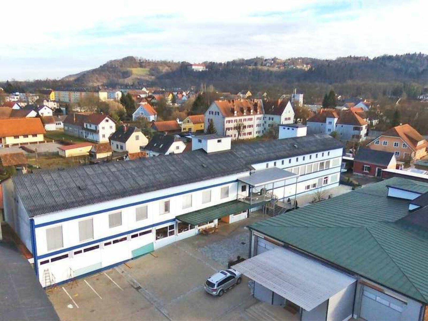 Luftaufnahme eines Gebäudekomplexes mit umliegenden Wohnhäusern und grünen Hügeln im Hintergrund.