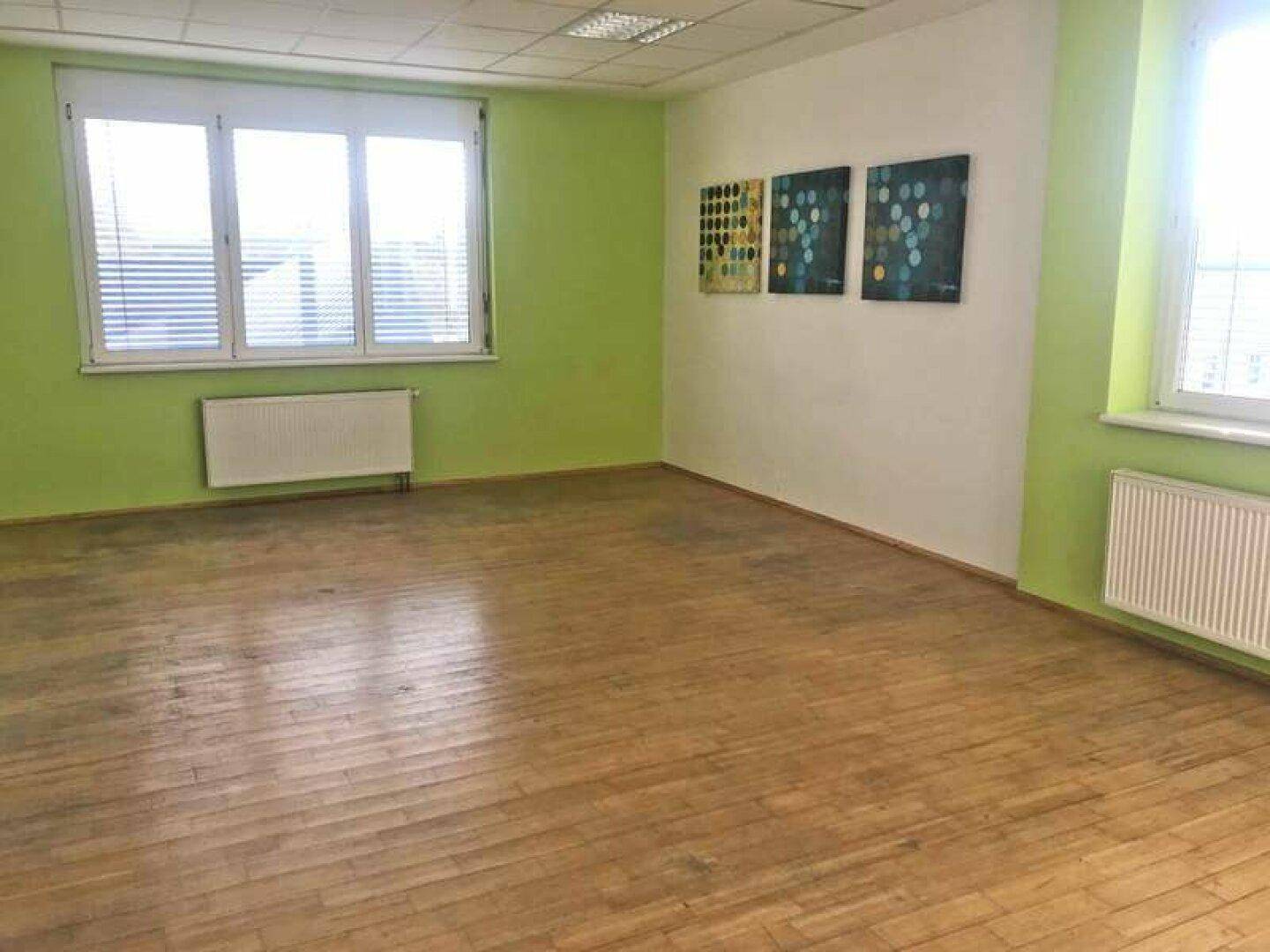 Leerer Schulungsraum mit hellgrünen Wänden, großen Fenstern und Holzboden, bereit für neue Einrichtung.