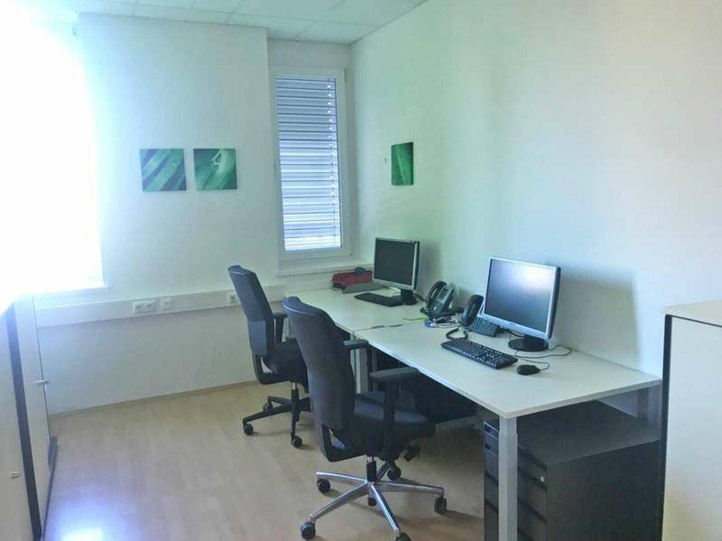 Helles Büro mit zwei Arbeitsplätzen, Computern und einem Fenster mit Jalousien für Privatsphäre.