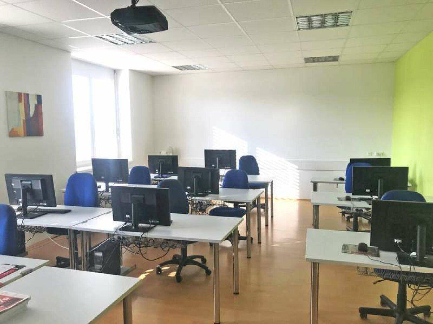 Schulungsraum mit mehreren Computern, Tischen und blauen Stühlen, ausgestattet für digitale Kurse.
