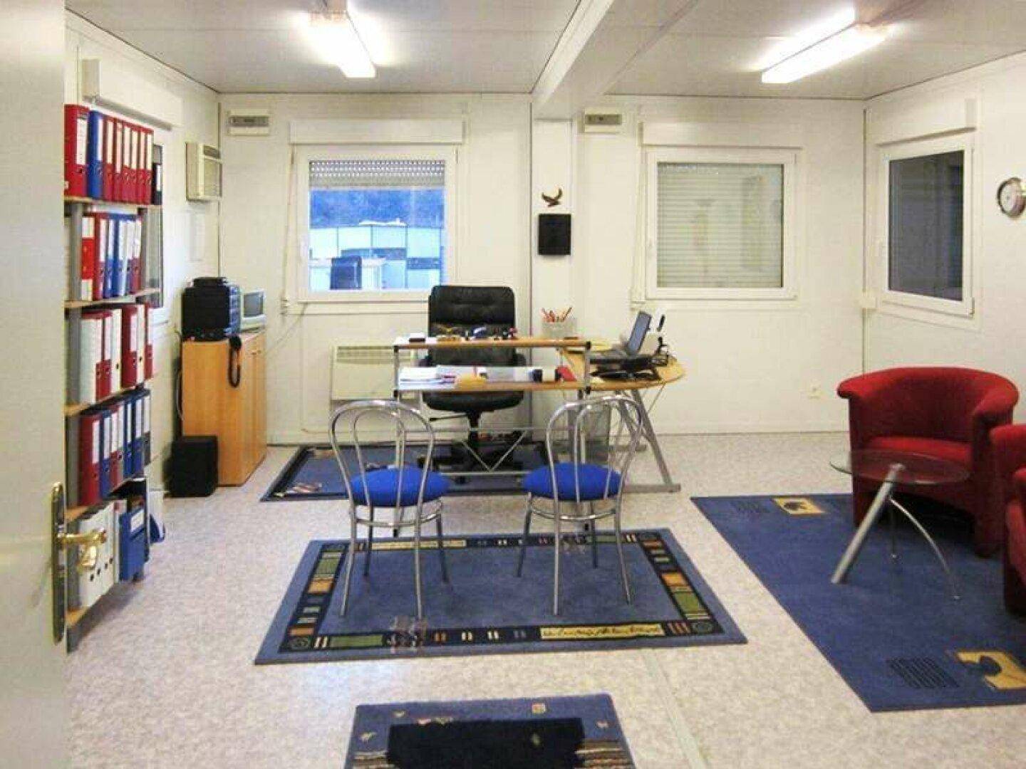 Funktionales Büro mit Schreibtisch, Bürostuhl, Regalen und bequemen roten Sesseln.