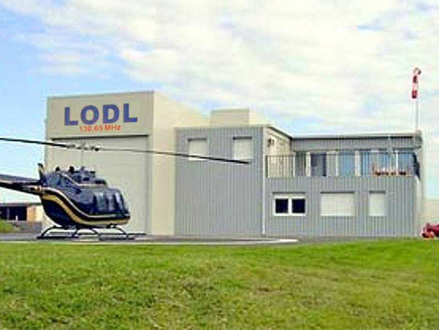 Außenansicht eines Gebäudes mit LODL-Beschriftung, einem Hubschrauber auf dem Landeplatz und einem Windsack.