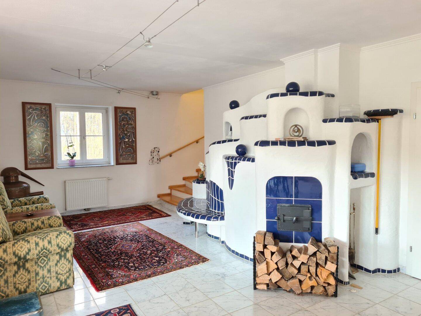 Geräumiger Wohnbereich mit Kachelofen, Holzstapel und traditionellen Teppichen auf Fliesenboden.