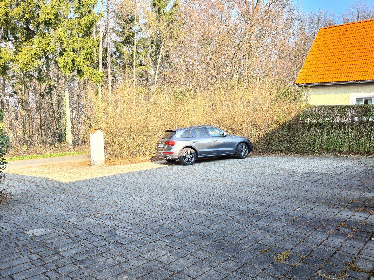 Großer gepflasterter Parkplatz vor dem Haus, umgeben von Bäumen und Sträuchern.