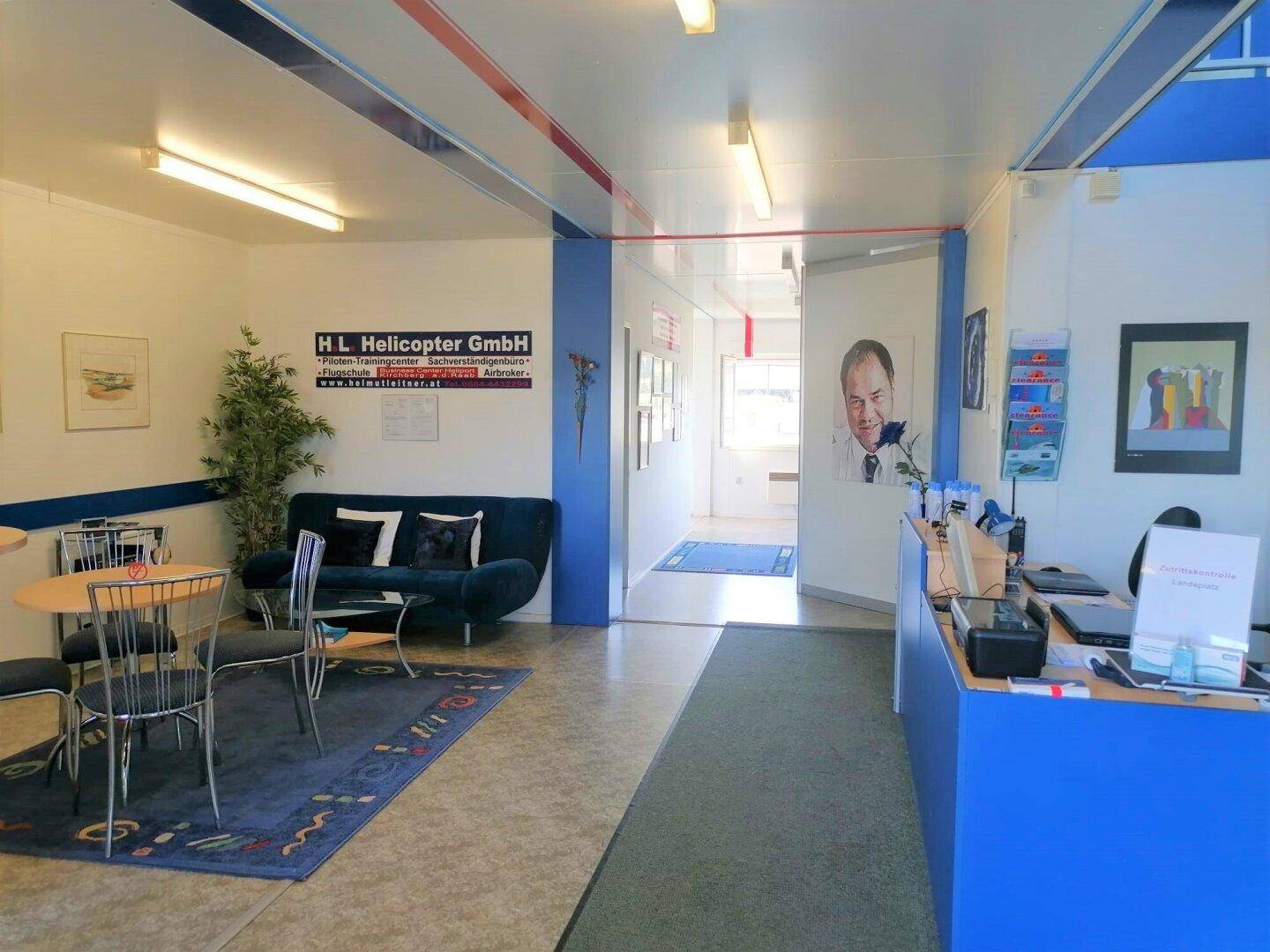 Einladender Empfangsbereich mit blauer Couch, kleinem Tisch, Rezeptionstheke und Zugang zu einem hellen Flur.