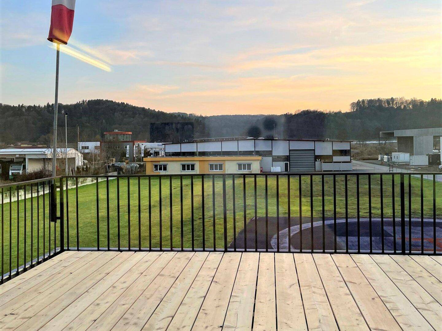 Blick von der Holzterrasse mit schwarzem Geländer auf eine grüne Landschaft und entfernte Gebäude bei Sonnenuntergang.