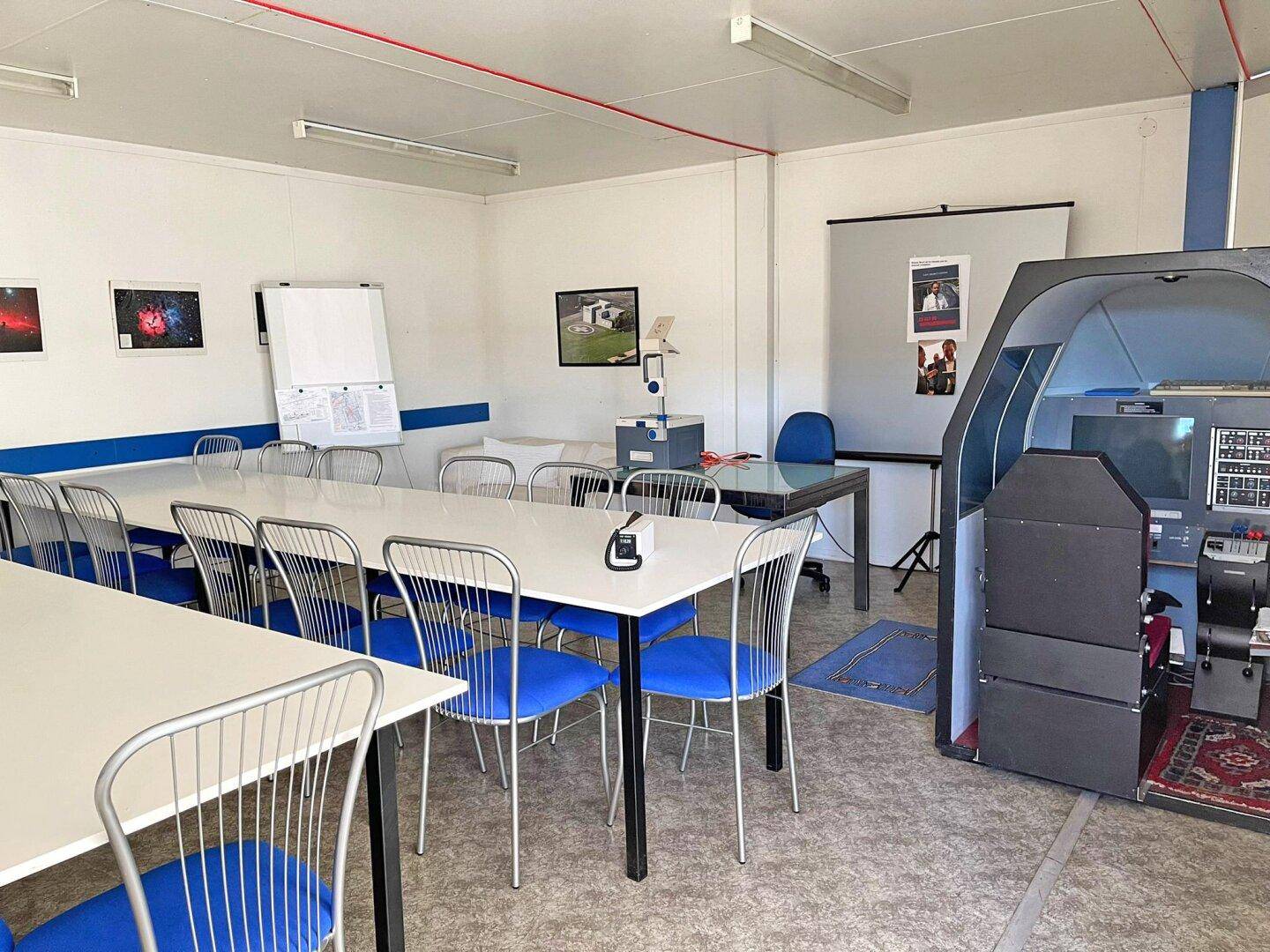 Heller Konferenzraum mit langen Tischen, blauen Stühlen, Whiteboard und einem Flugsimulator für Schulungszwecke.