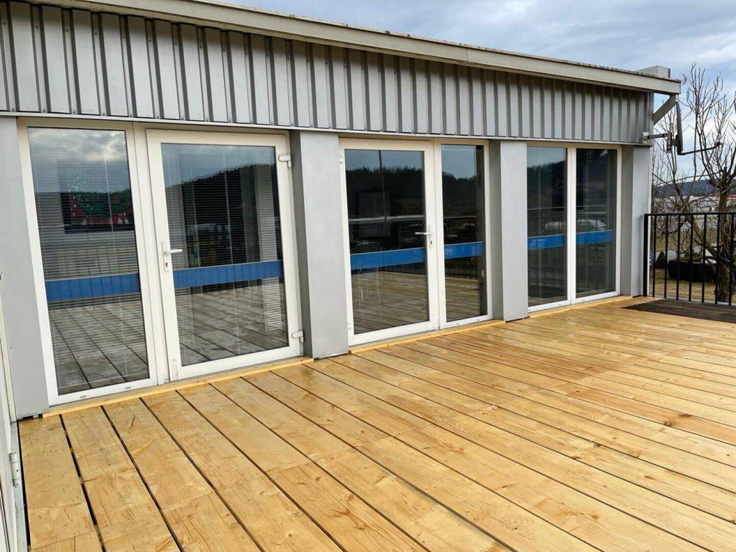Geräumige Holzterrasse mit mehreren Glastüren, die Zugang zum Gebäude bieten und einen gepflegten Eindruck machen.