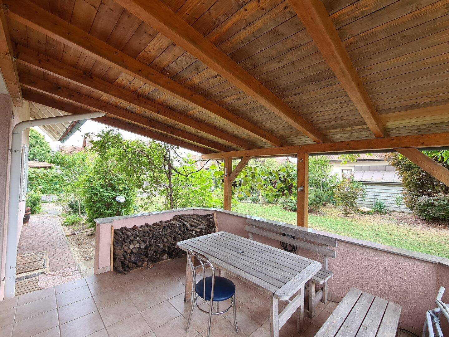 Überdachte Terrasse mit Holzdach, Fliesenboden und Sitzgelegenheiten im Freien.