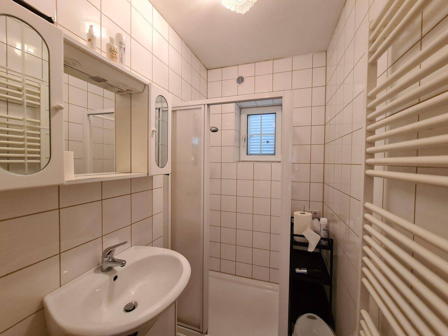 Kleines Badezimmer mit Dusche, Waschbecken, Fenster und beheiztem Handtuchhalter.