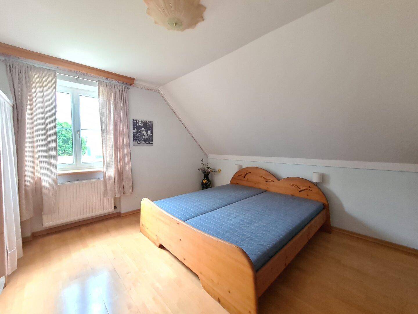 Helles Schlafzimmer mit Holzbett, Parkettboden und Fenster mit Blick ins Grüne.
