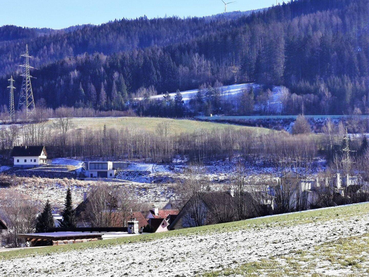 Weitläufige Landschaft mit Wohnhäusern, bewaldeten Hügeln und schneebedeckten Flächen im Winter.