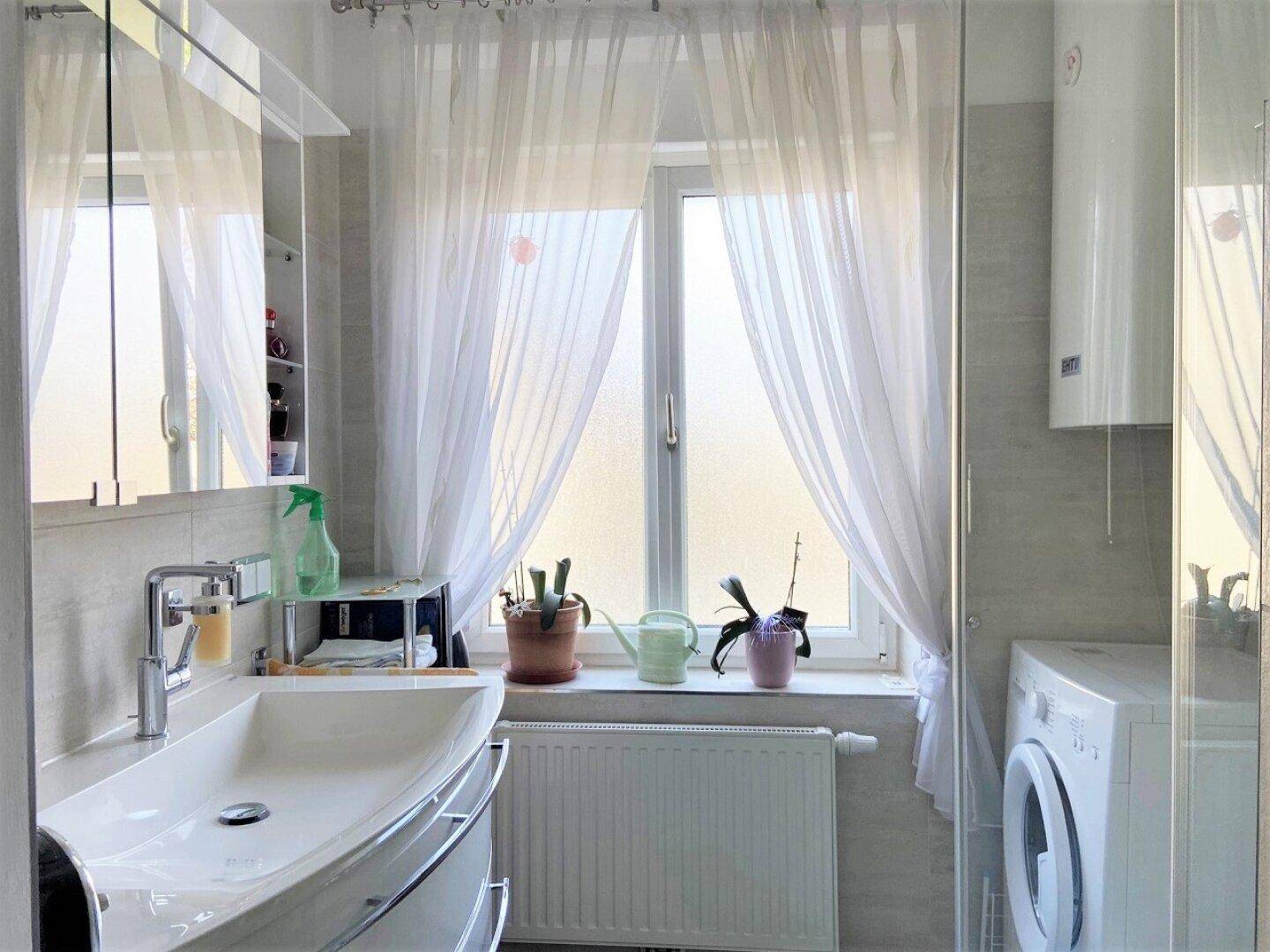 Helles Badezimmer mit Waschbecken, Fenster und Waschmaschine, ausgestattet mit einer Duschkabine und Fliesenboden.
