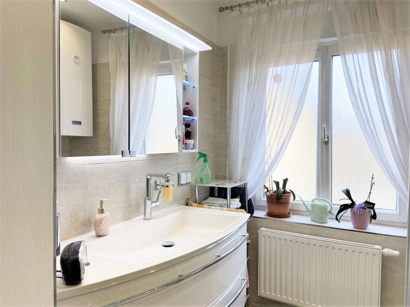 Helles Badezimmer mit modernem Waschtisch, Spiegelschrank und Fenster mit Vorhängen.