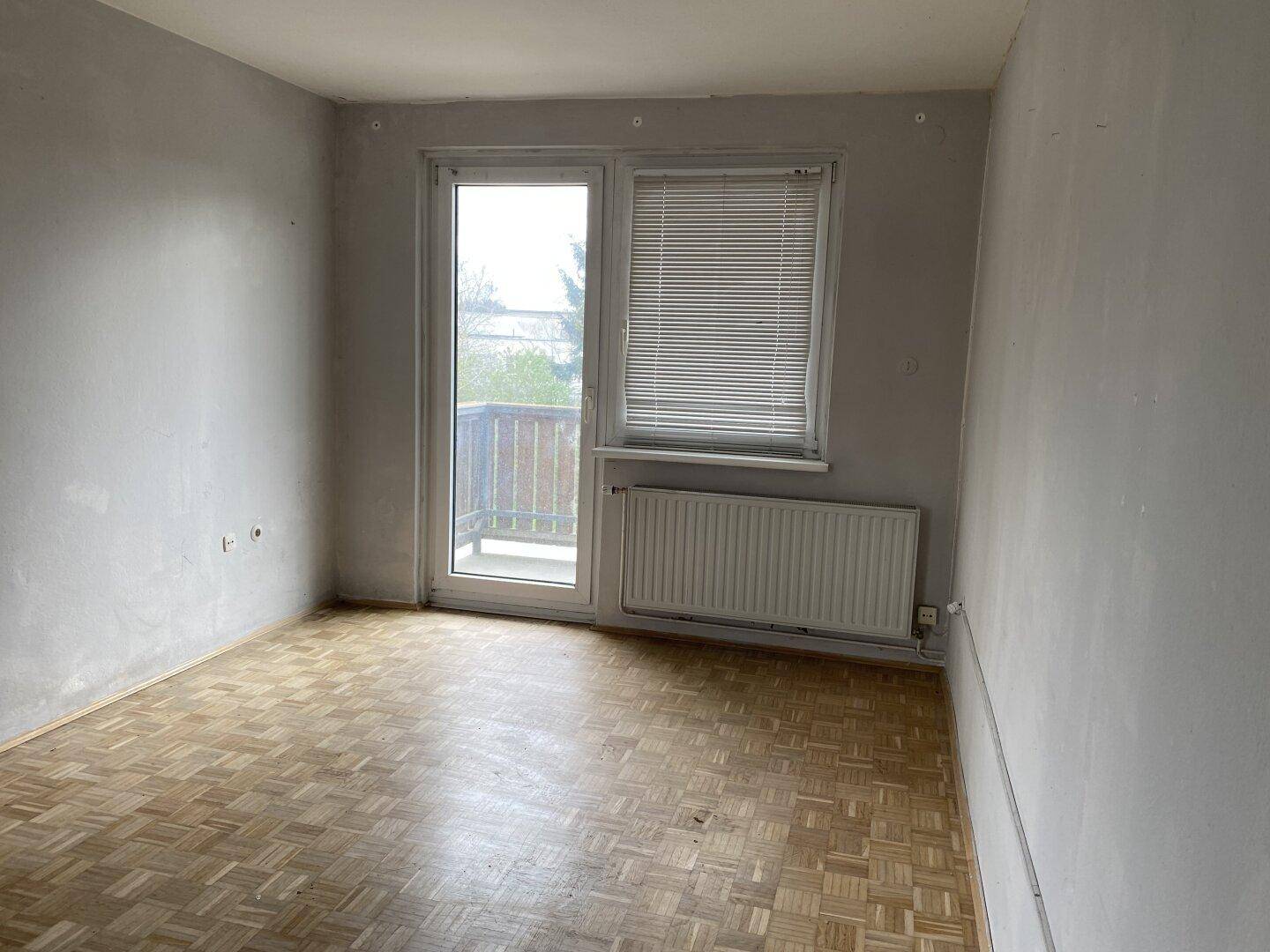 Leeres Zimmer mit hellem Parkettboden, grauen Wänden und Zugang zu einem Balkon.