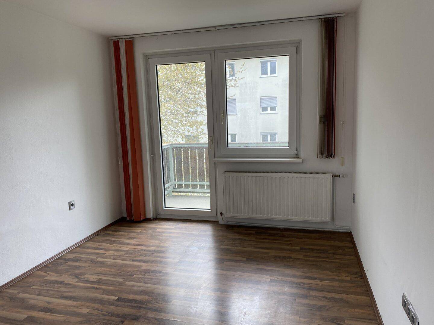 Helles Zimmer mit Laminatboden, Heizkörper und Zugang zu einem Balkon mit Blick auf ein Gebäude.