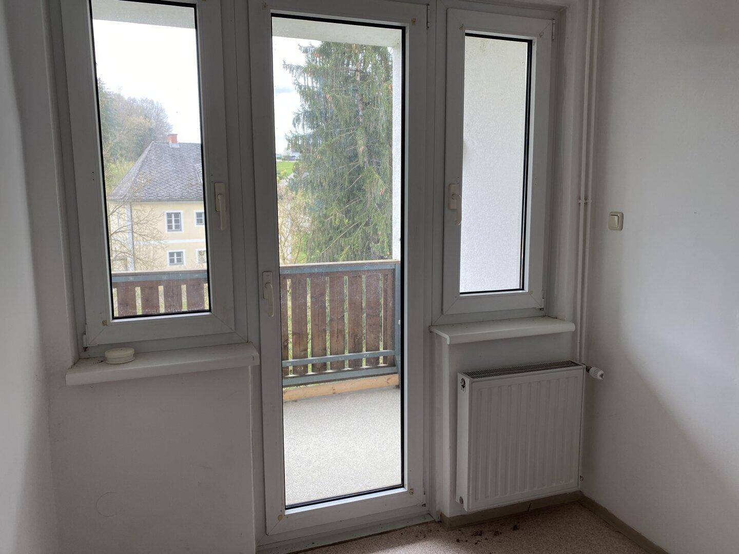 Heller Raum mit großen Fenstern, Heizkörper und Zugang zu einem Balkon mit Blick ins Grüne.