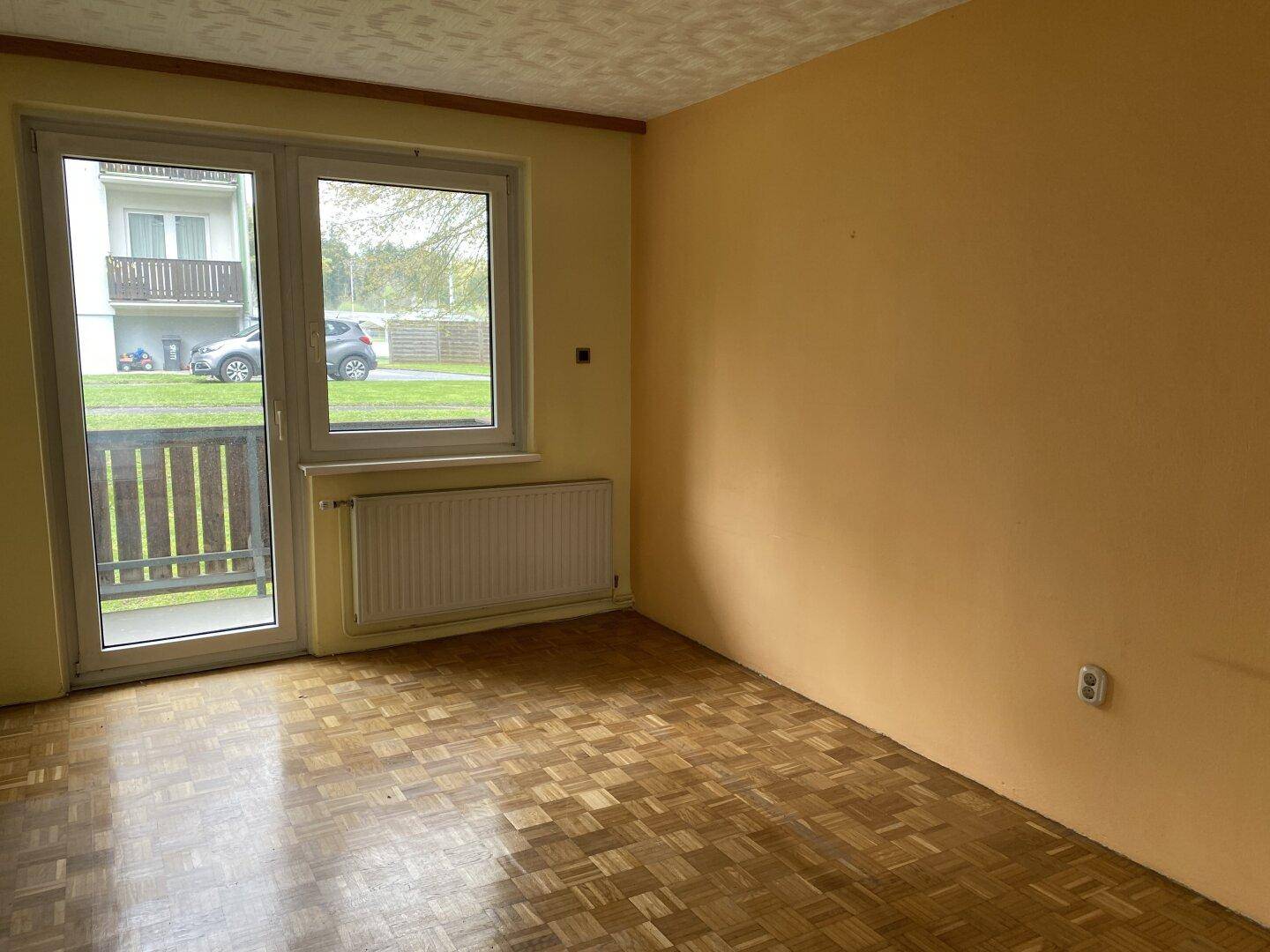 Geräumiges Zimmer mit gelben Wänden, Parkettboden und Balkonzugang.