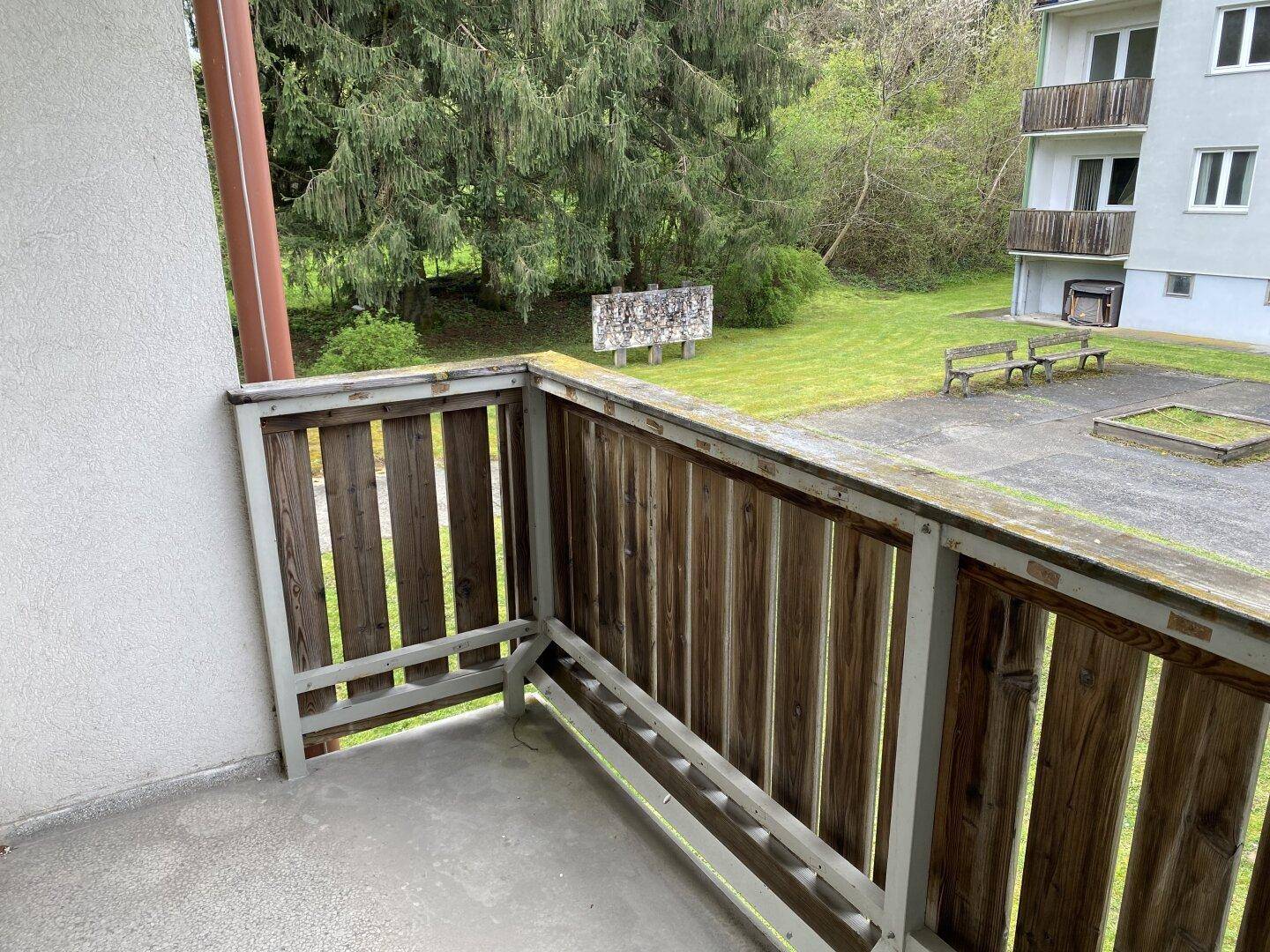 Balkon mit Holzbalkonbrüstung und Blick auf eine Grünfläche mit Bäumen und ein Nachbargebäude.
