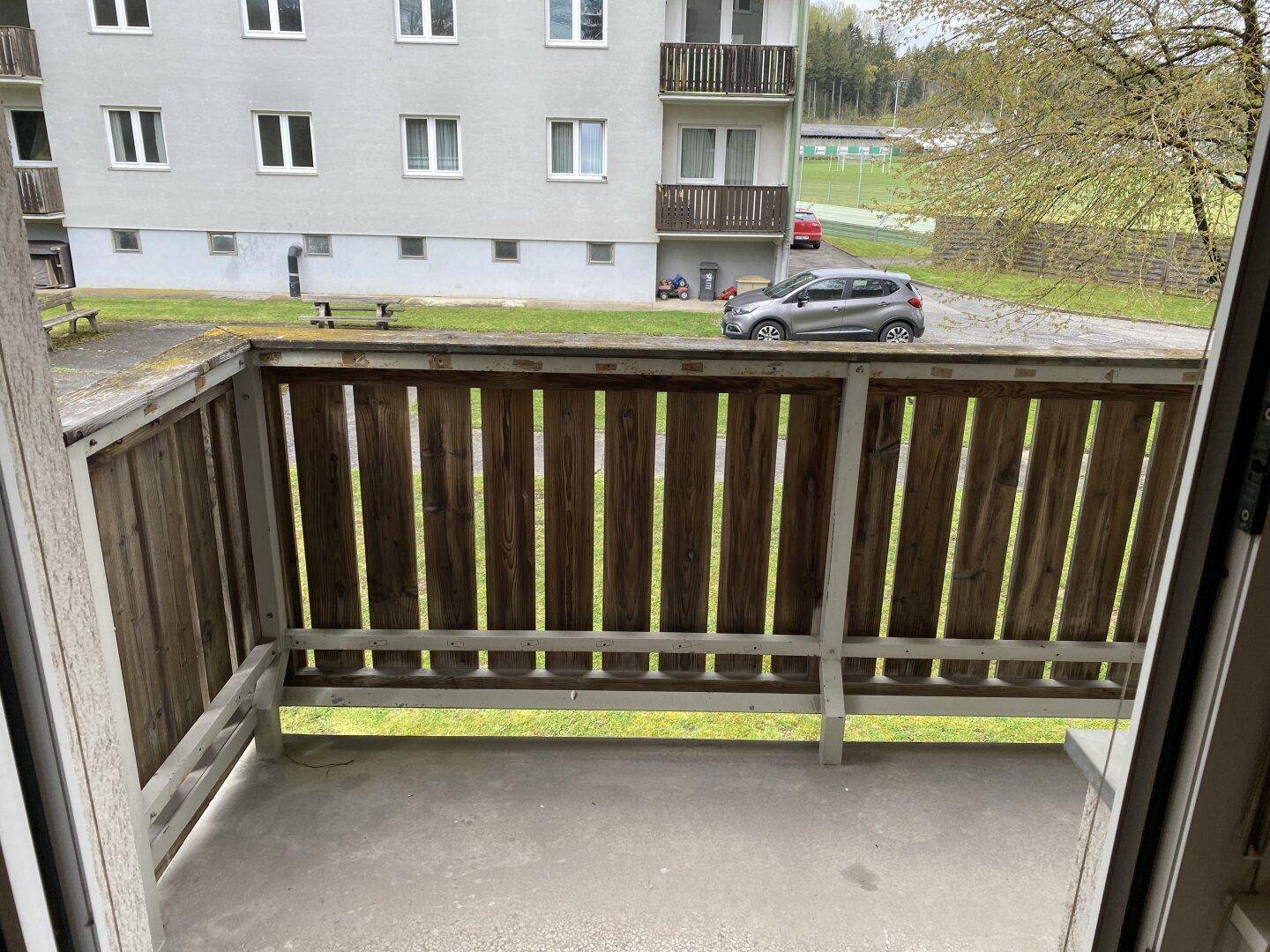 Balkon mit Holzbalkonbrüstung und Blick auf eine Grünfläche mit Parkplatz und Wohngebäude.