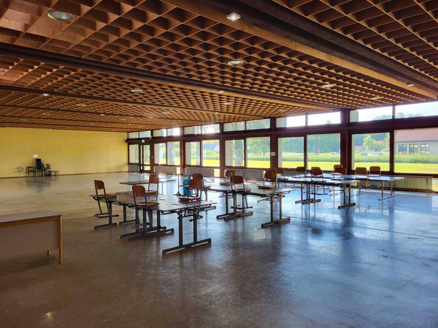Geräumiger Mehrzweckraum mit Tischen und Stühlen, Holzgitterdecke und weitem Blick ins Grüne durch die Fensterfront.