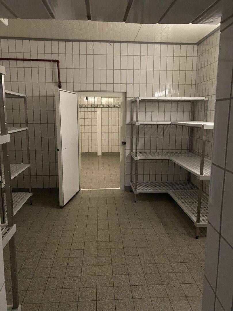 Gefliester Lagerraum mit Metallregalen und einer Tür, die zu einem weiteren gefliesten Bereich führt.