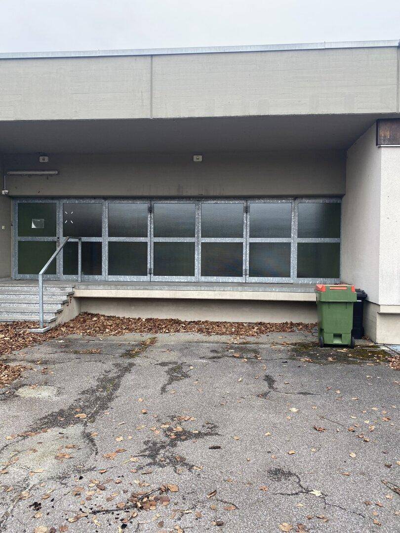 Außenansicht eines Gebäudes mit großen, vergitterten Fenstern, einer Laderampe und einem asphaltierten Vorplatz.