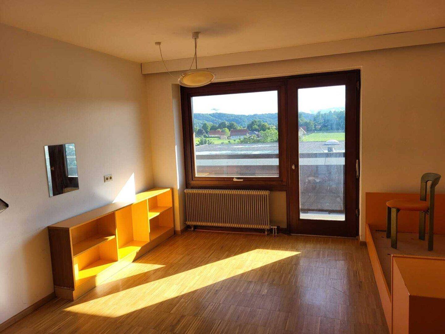 Sonniges Zimmer mit großem Fenster, Blick ins Grüne und Zugang zu einem Balkon oder Terrasse.