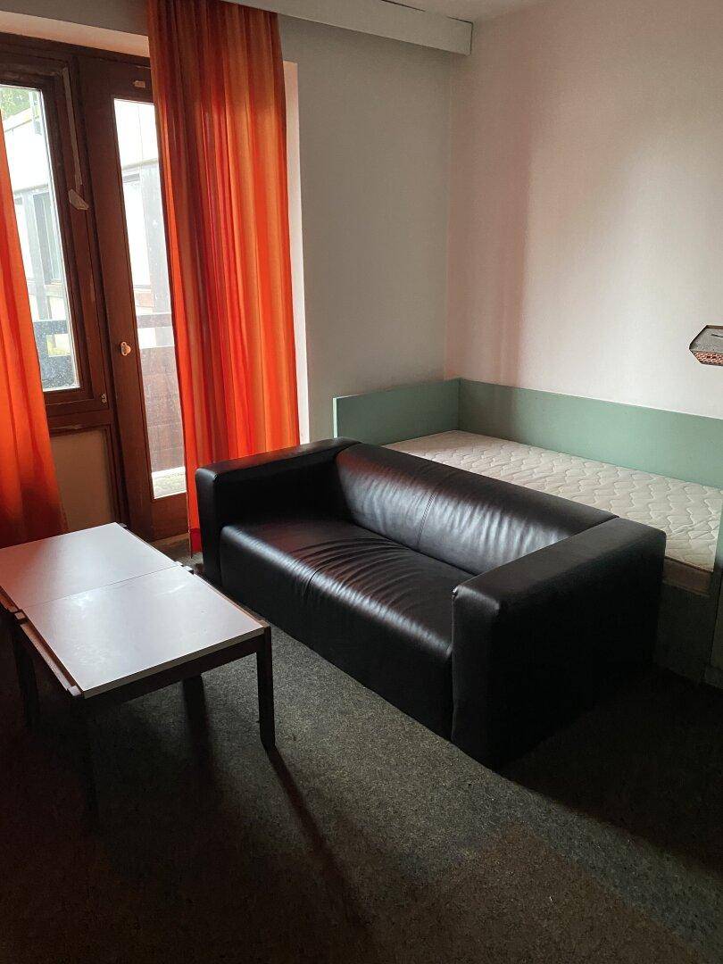 Gemütliches Zimmer mit schwarzem Sofa, kleinem Tisch und Zugang zu einem Balkon oder Terrasse.