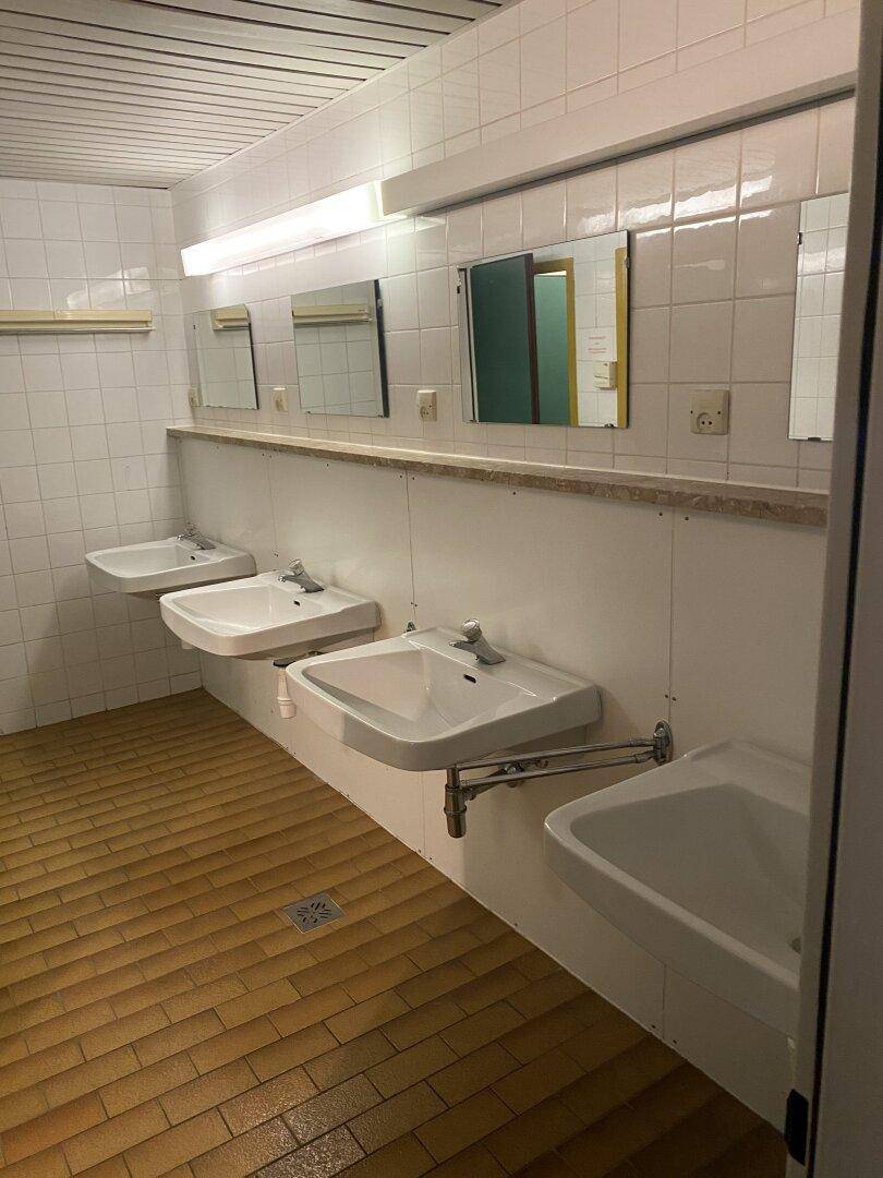 Funktionales Gemeinschaftsbad mit mehreren Waschbecken, weißen Fliesen und braunem Fliesenboden.