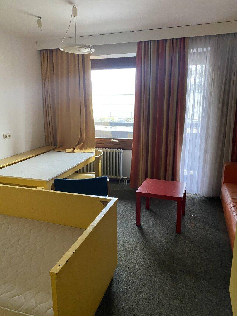 Zimmer mit mehreren Betten, kleinem roten Tisch und Fenster mit Blick nach draußen.