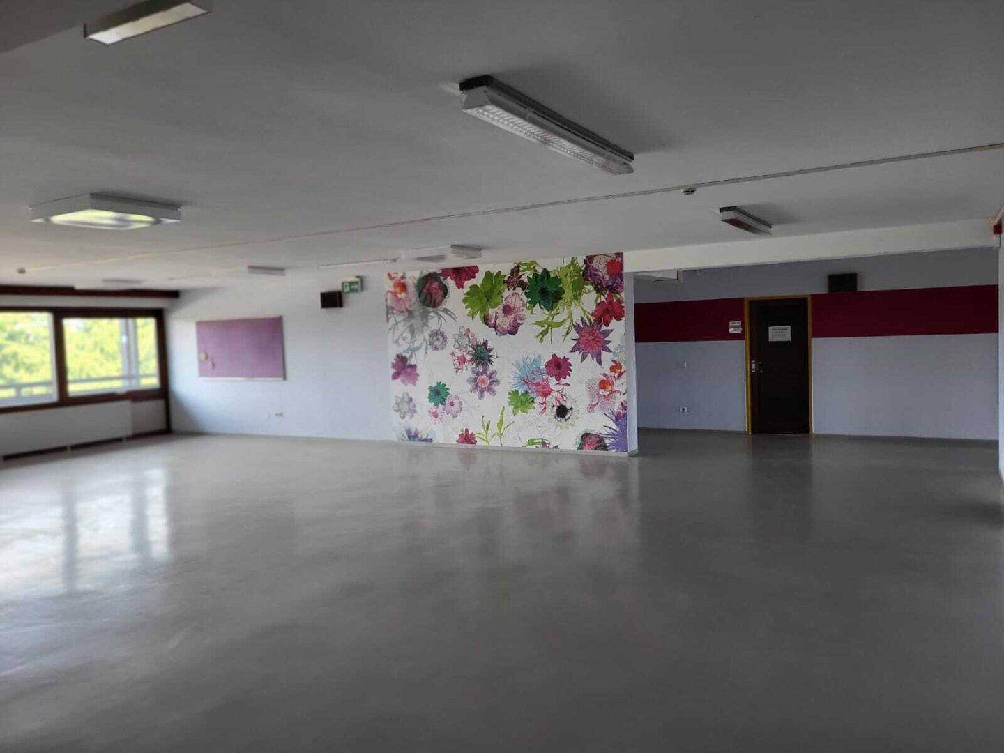 Großer, leerer Mehrzweckraum mit floraler Wanddekoration, großen Fenstern und glattem Bodenbelag.