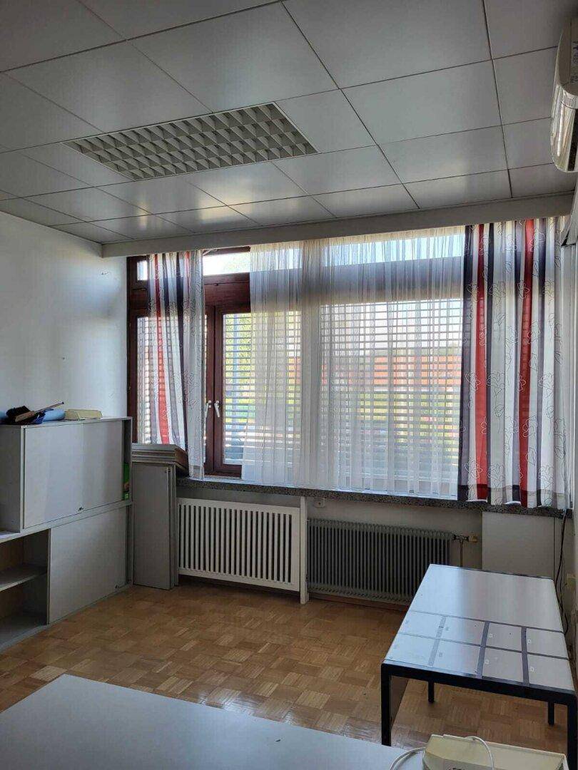 Kleines Büro mit Holzparkett, Fenster mit Jalousien und einem Heizkörper unter dem Fenster.