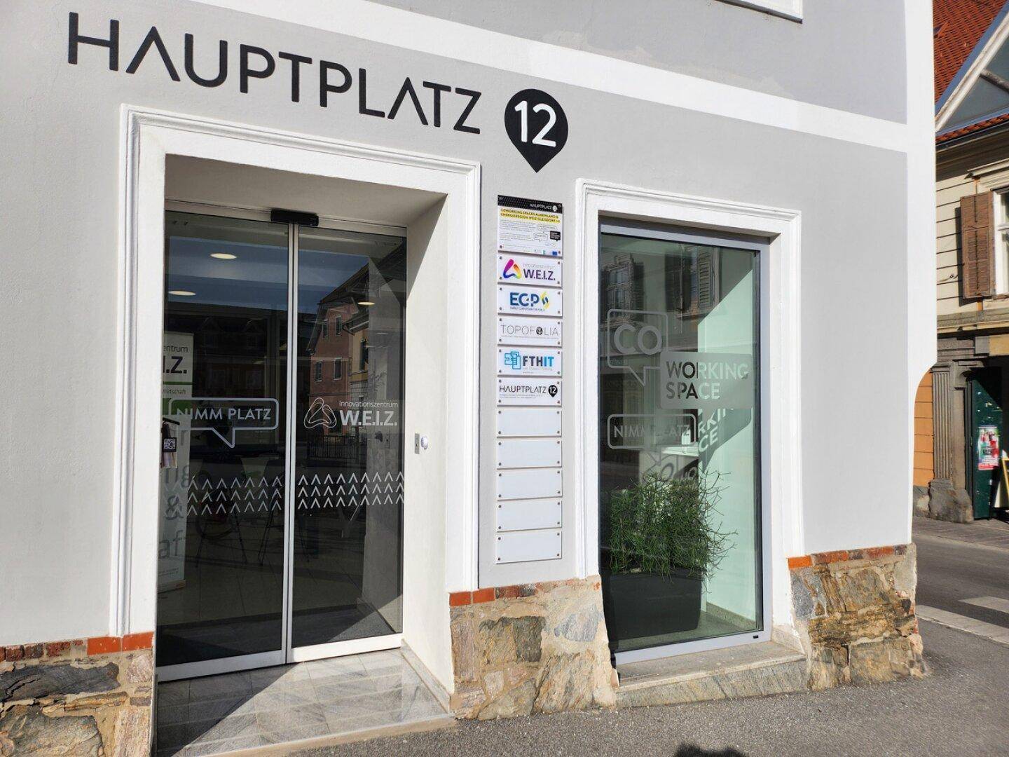 Moderner Eingangsbereich am Hauptplatz 12 mit automatischen Glastüren und einer hellgrauen Fassade.