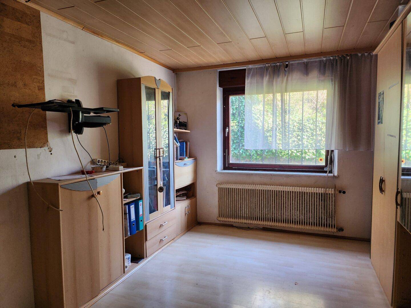 Zimmer mit Holzdecke, Laminatboden und einem Heizkörper unter dem Fenster.