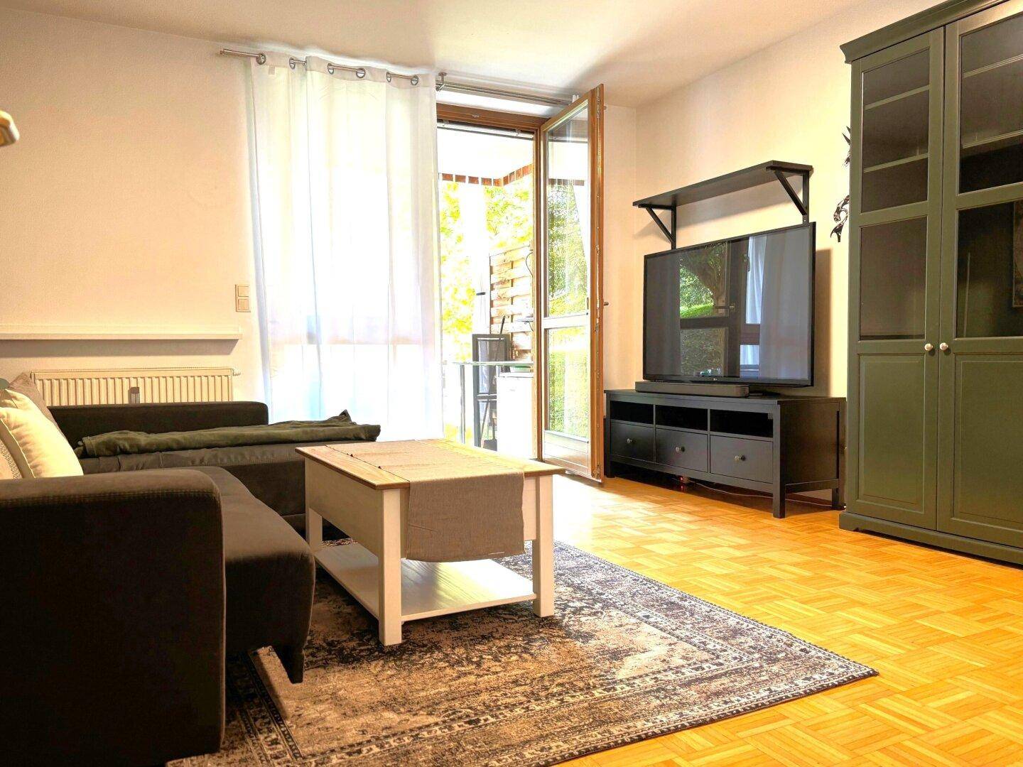 Geräumiges Wohnzimmer mit Fernseher, Schrank und Zugang zu einem sonnigen Balkon.