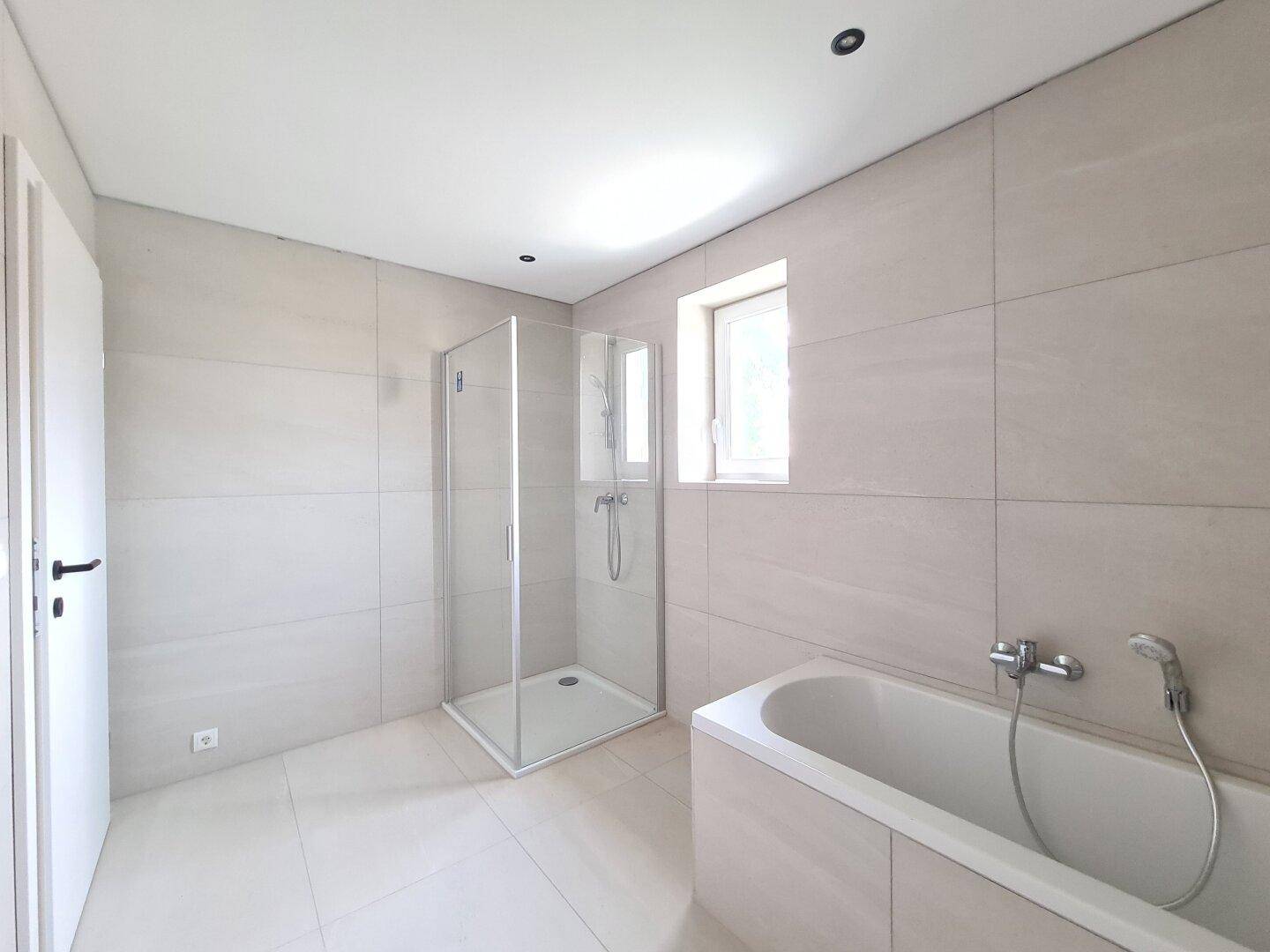 Modernes Badezimmer mit Badewanne, Duschkabine und Fenster, in hellen Fliesen gehalten.