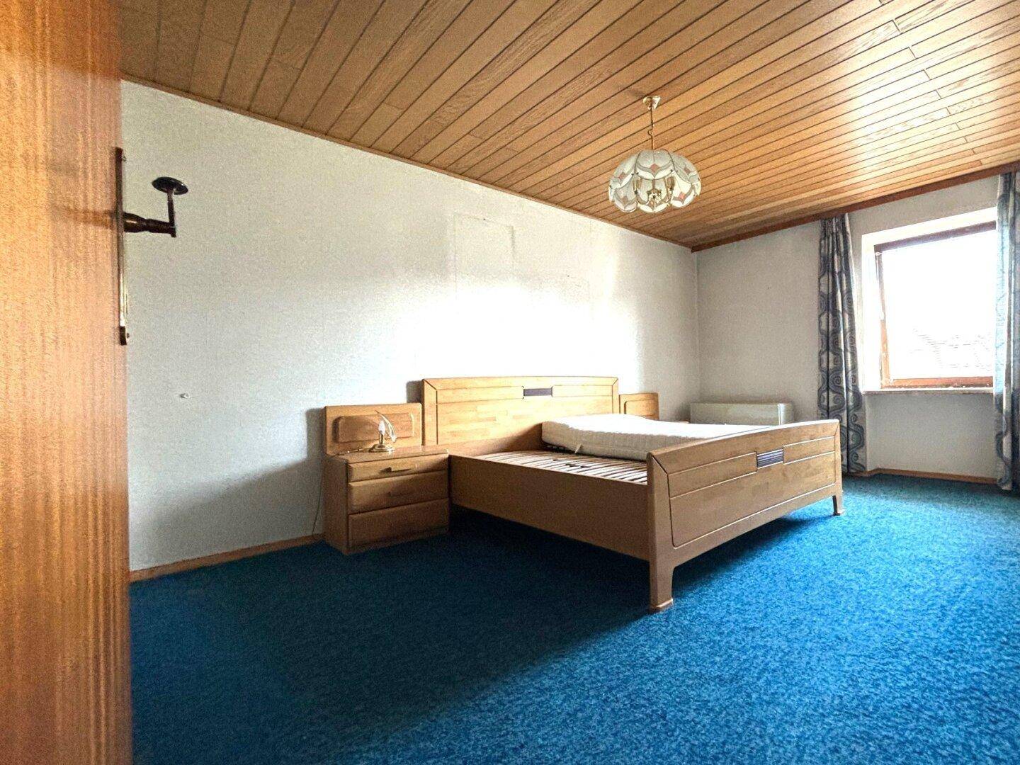 Schlafzimmer mit blauem Teppichboden, Holzdecke und einem einfachen Holzbett.