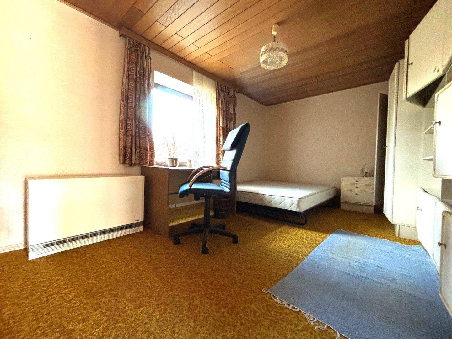 Helles Zimmer mit gelbem Teppichboden, Holzdecke und einem Schreibtisch am Fenster.