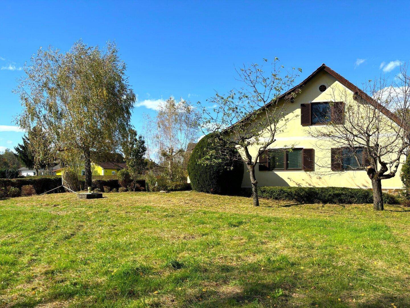 Seitenansicht des Hauses mit großem Rasen, Bäumen und Sträuchern unter blauem Himmel.