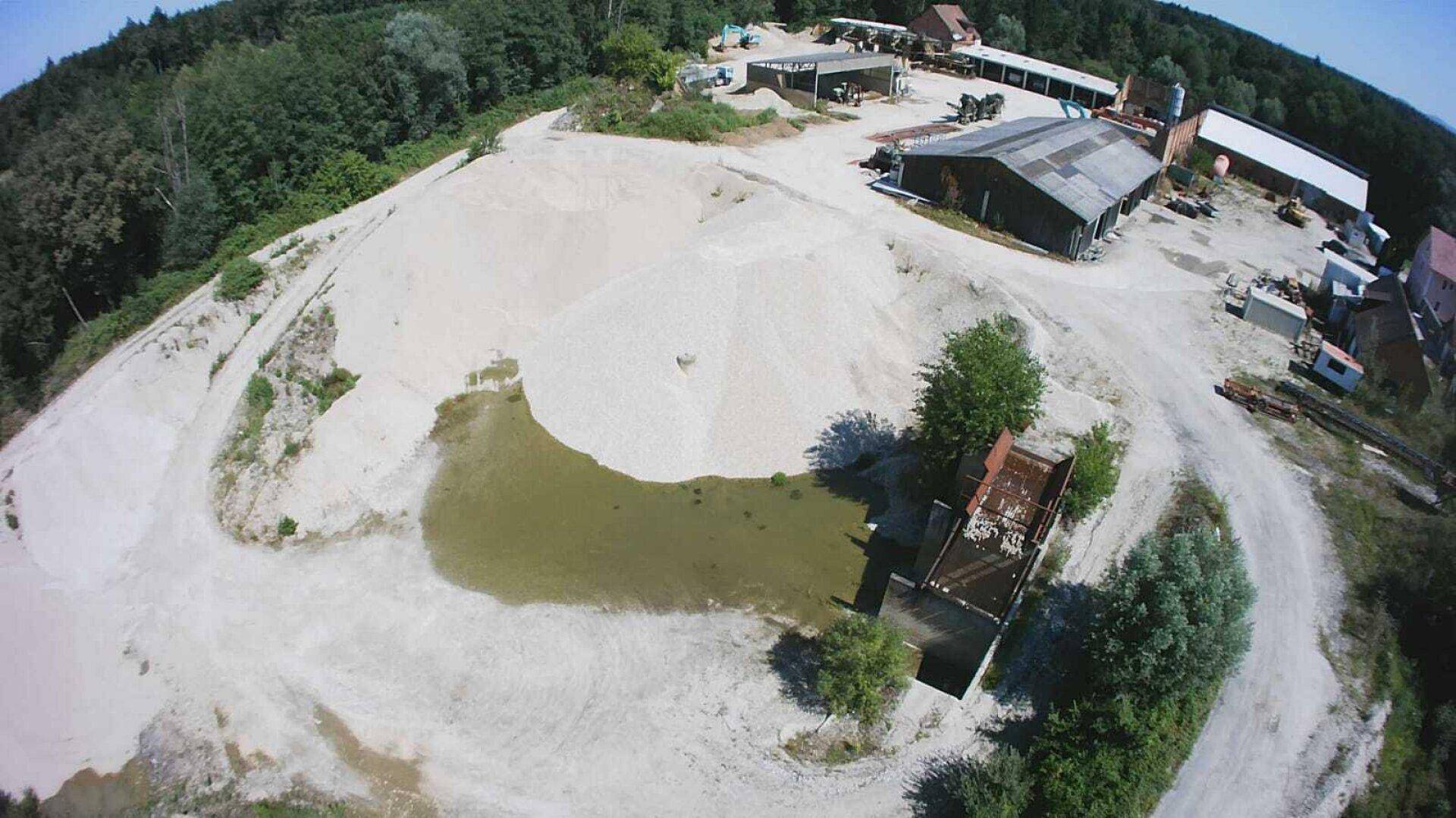 Luftaufnahme von riesigen Sand- und Kieshaufen mit einem kleinen Teich und Betriebsgebäuden im Hintergrund.