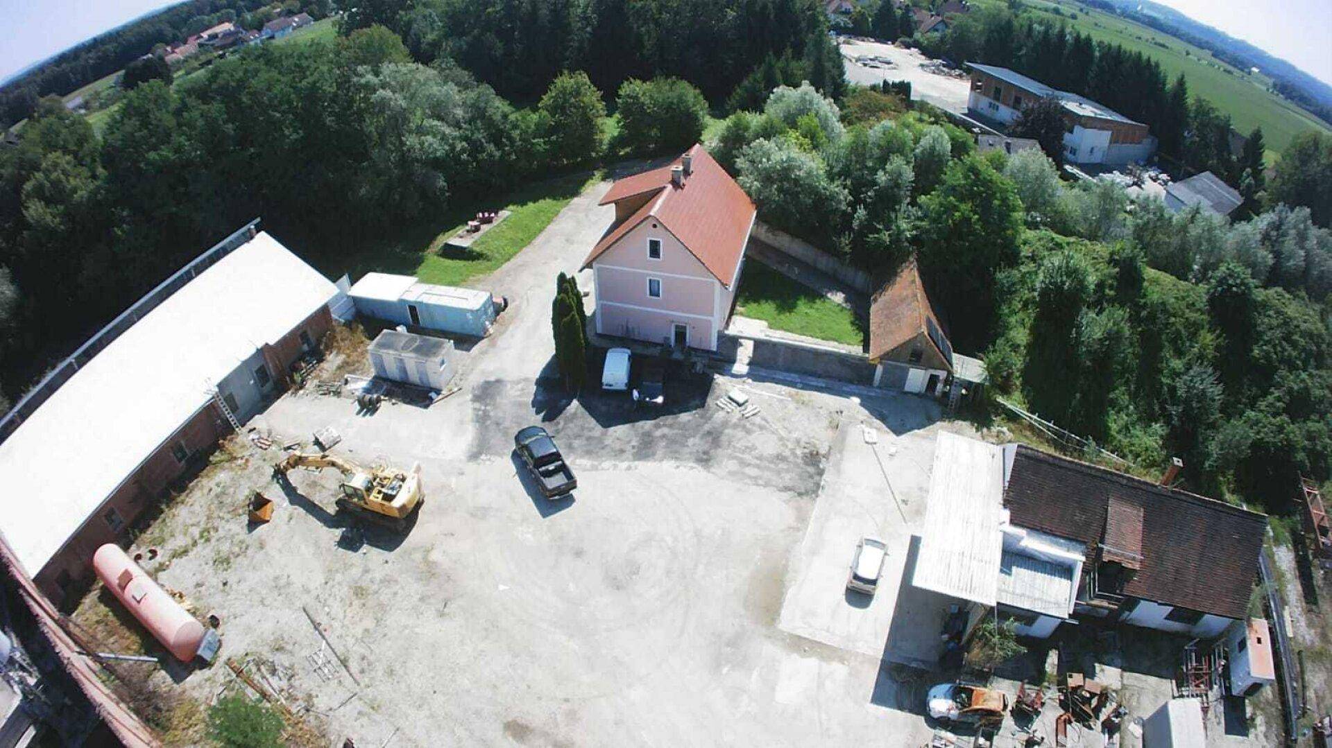Luftaufnahme eines Wohnhauses mit rotem Dach neben dem Betriebsgelände, umgeben von Bäumen und Parkplätzen.
