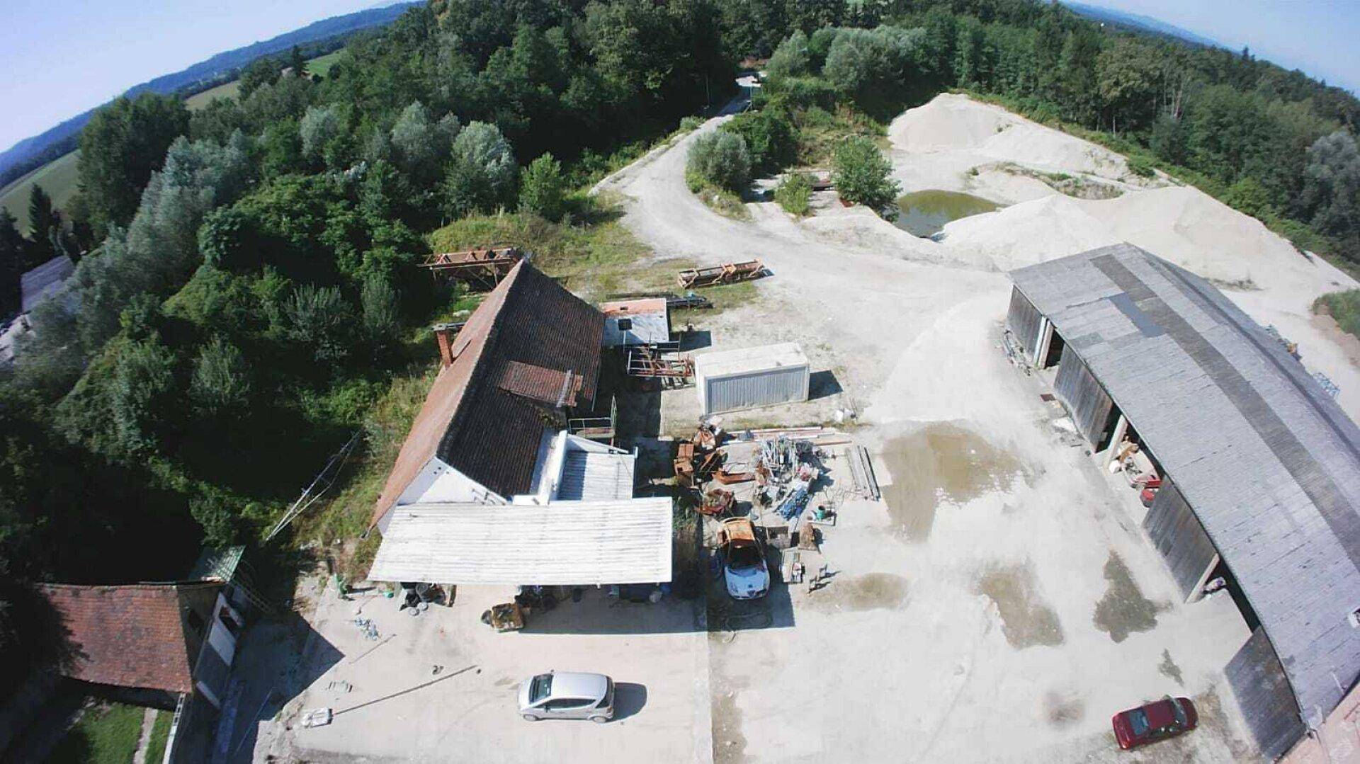 Blick von oben auf die Industriegebäude, Materiallager und die angrenzenden Waldgebiete unter blauem Himmel.
