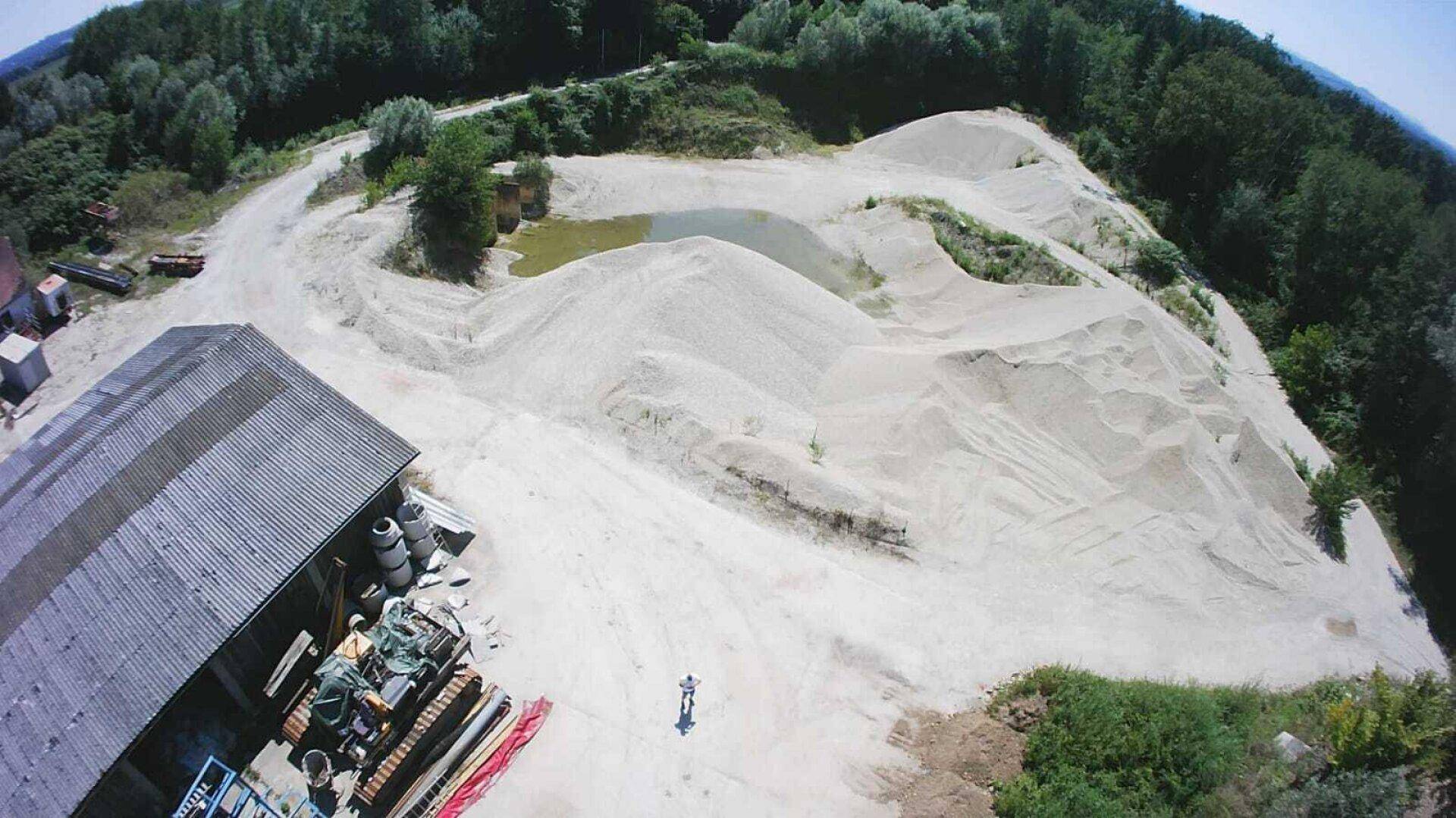 Luftaufnahme von ausgedehnten Sandhügeln mit einem kleinen Teich und einem Gebäude am Horizont.
