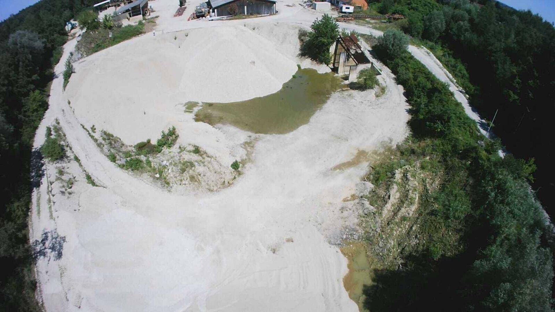 Weitläufige Sand- und Kieslandschaft aus der Vogelperspektive mit einem kleinen Teich und einem Nebengebäude.