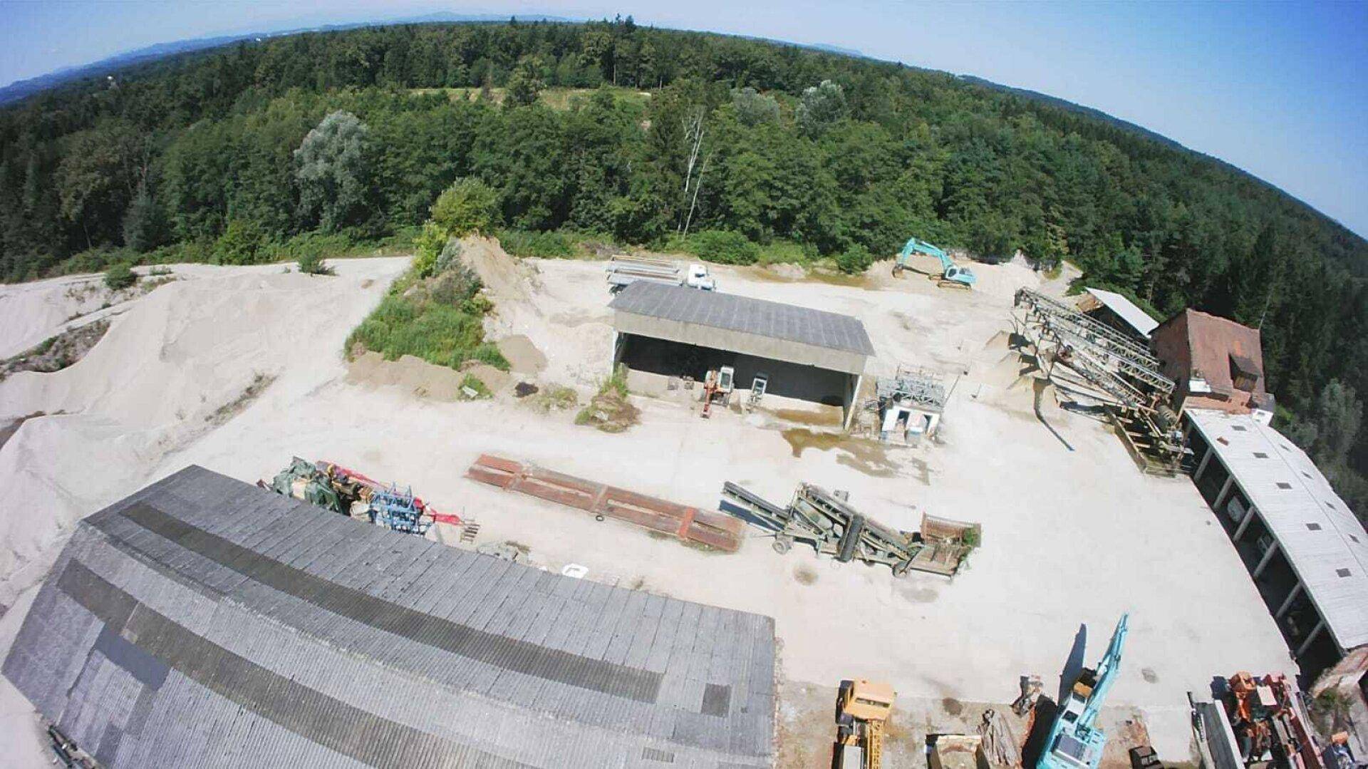 Blick von oben auf das Betriebsgelände mit Lagerhallen, Maschinen und Materialhaufen, umgeben von Wald.