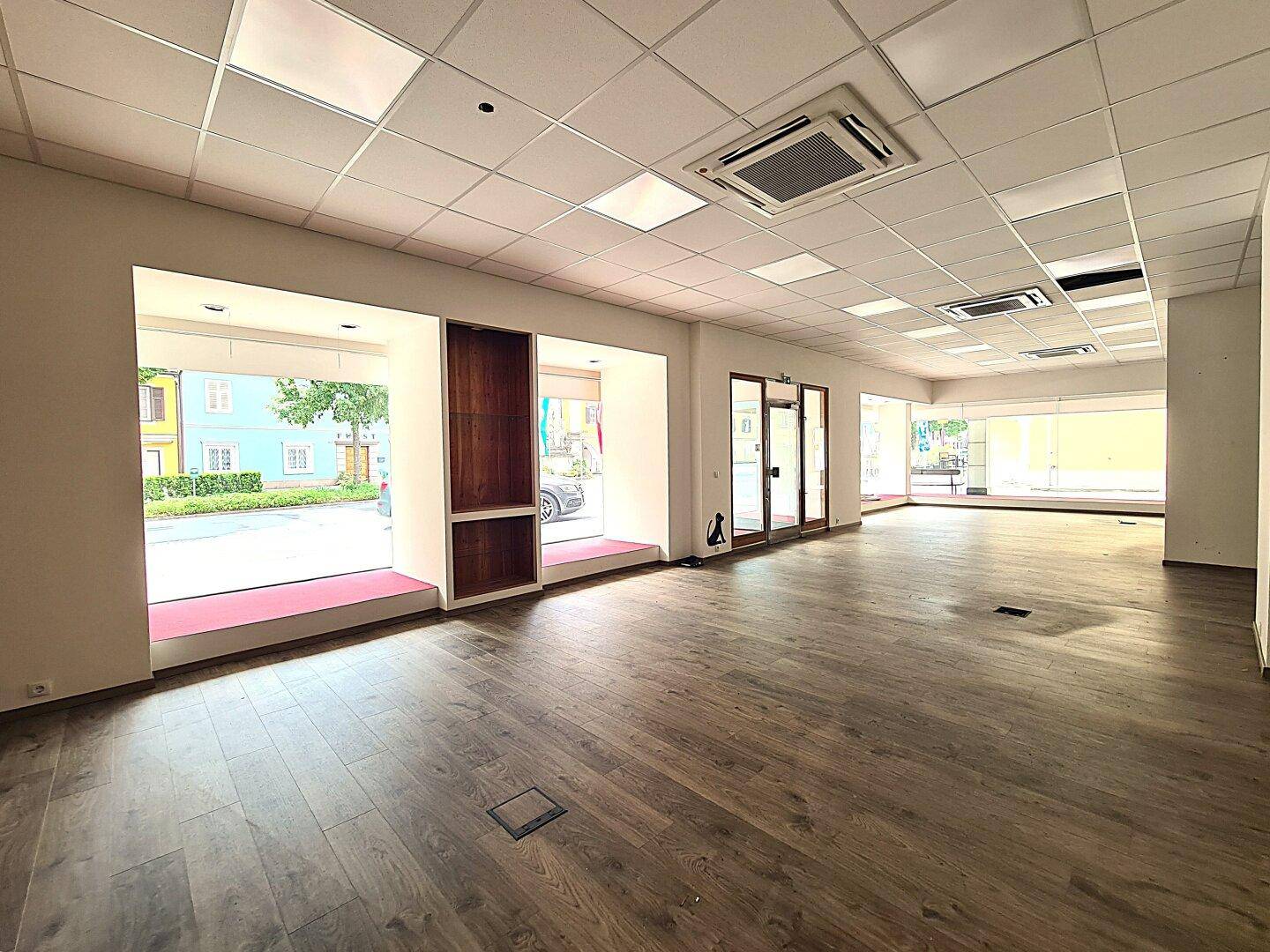 Geräumiger, leerer Geschäftsraum mit Laminatboden, abgehängter Decke und großen Schaufenstern zur Straße.